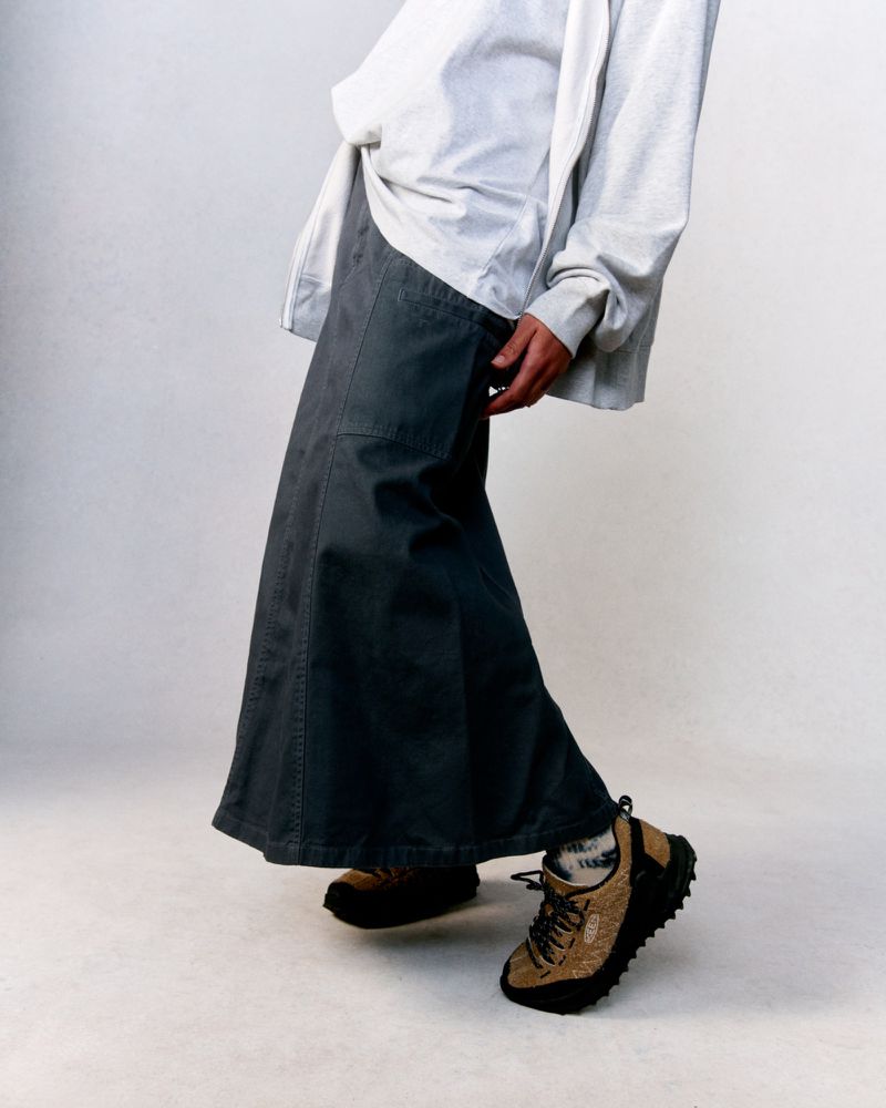 Person wears grey long-sleeved shirt, long denim skirt, tan and black athletic shoes in a fashion studio shot, against a light backdrop.