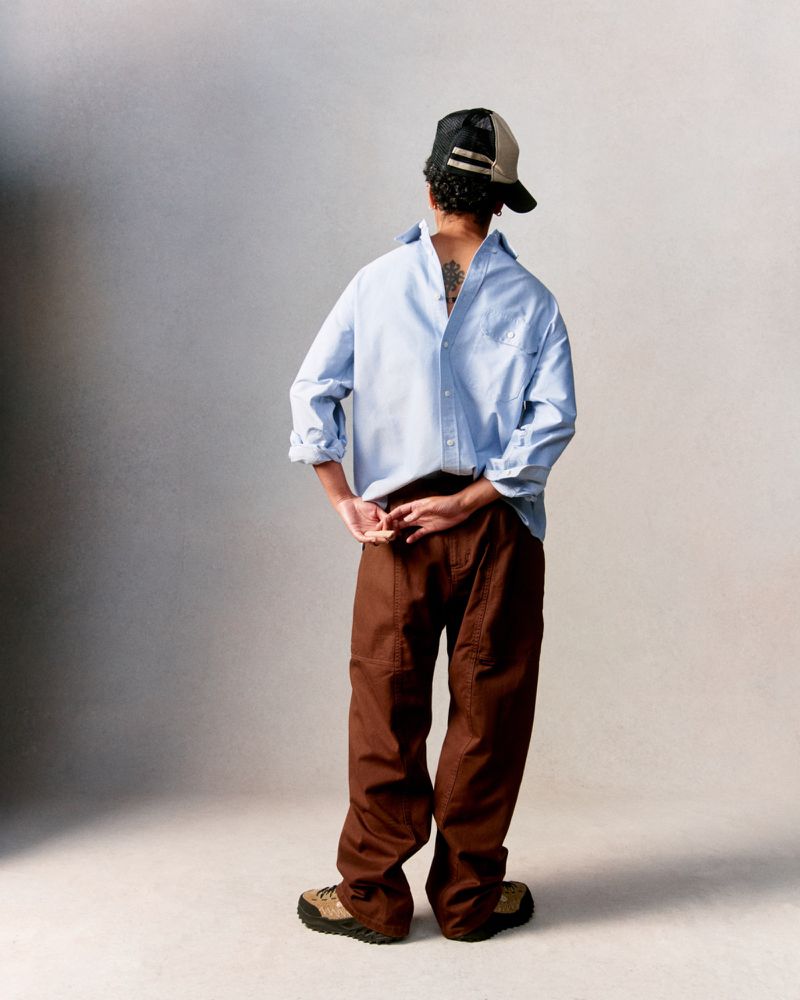 Man in casual attire: light blue shirt, brown pants, and baseball cap, captured from behind in a full shot against a neutral backdrop.