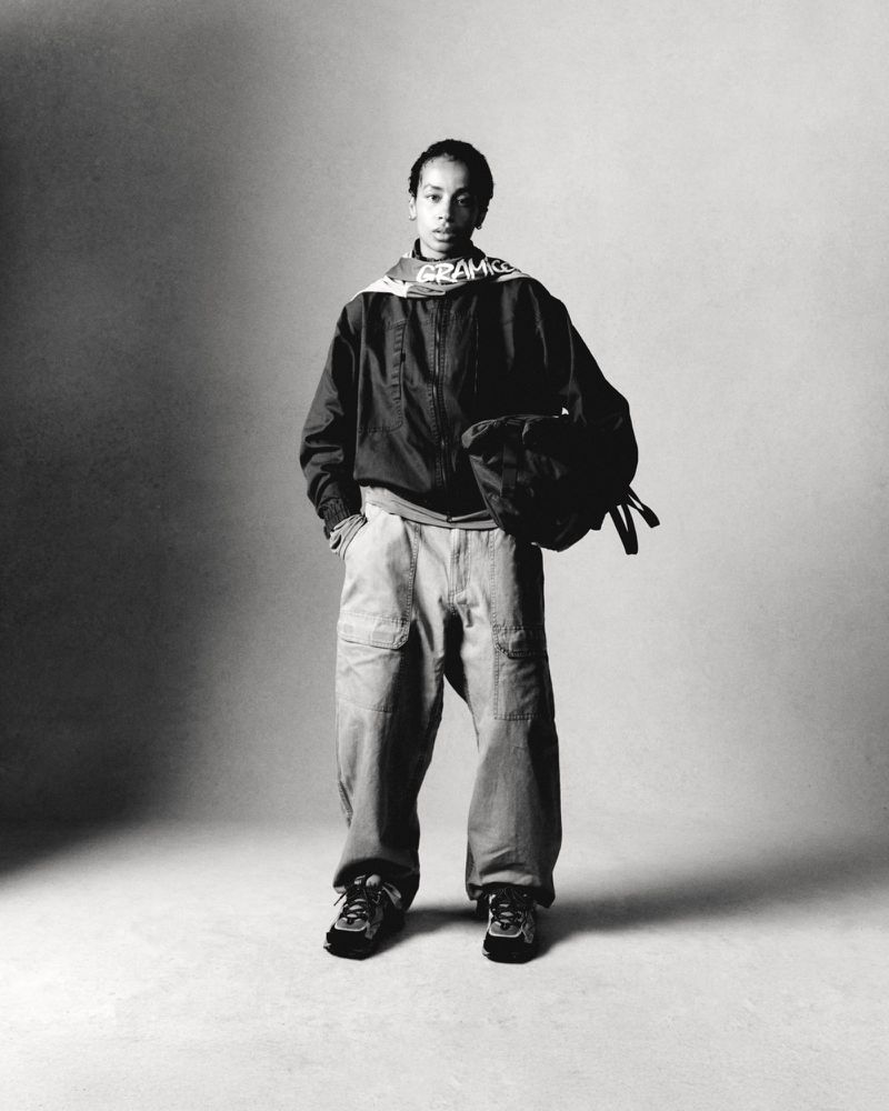 Monochrome shot of a young adult in casual athletic wear, featuring an unzipped jacket, baggy pants, sneakers, and carrying a bag.
