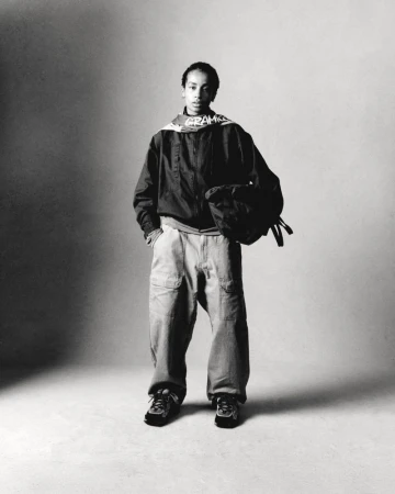 Monochrome shot of a young adult in casual athletic wear, featuring an unzipped jacket, baggy pants, sneakers, and carrying a bag.