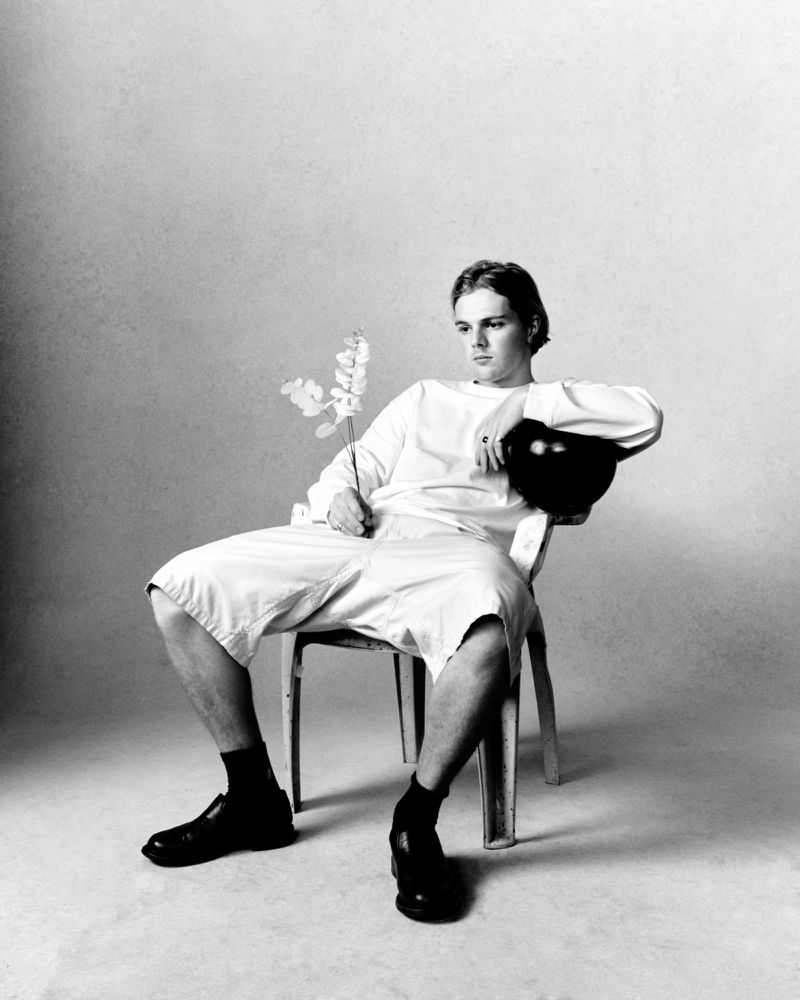Monochrome portrait of a young adult male sitting on a chair, holding a flower, with a dark helmet resting on the chair.
