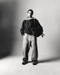 Model standing on a seamless studio floor wearing a dark jacket, scarf, and loose trousers, holding a bag.