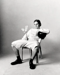 Young man seated on a chair holding a flower against a plain light studio background