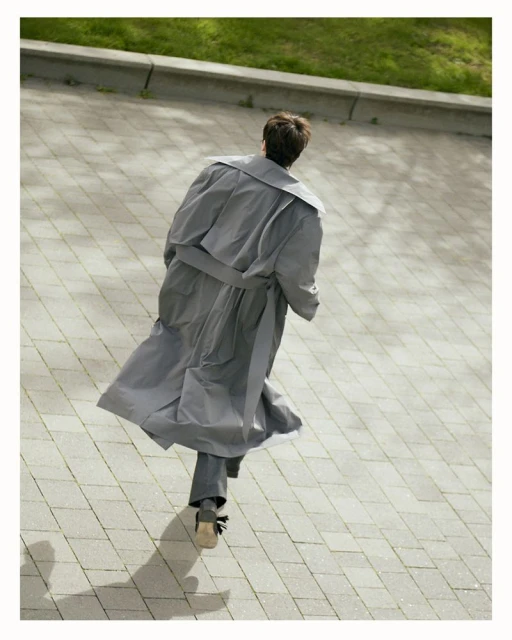 A person walks on a paved surface in a gray overcoat and trousers. A hint of green plants is visible in the background.