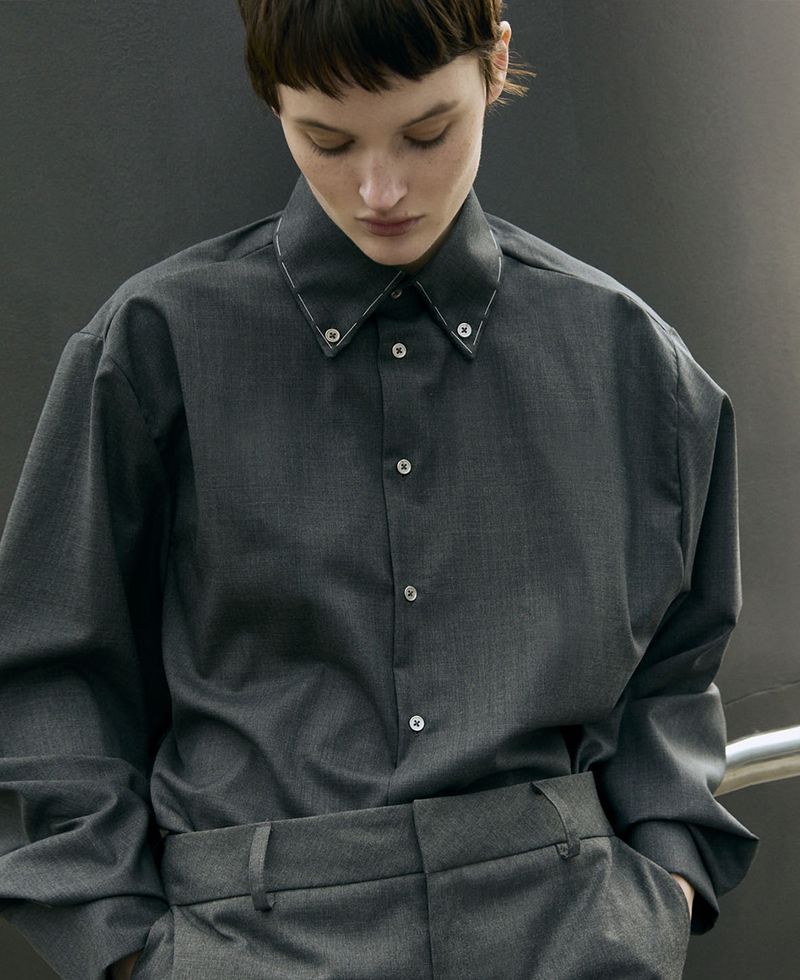 Portrait of a woman in a gray button-down shirt and matching trousers against a gray wall, looking down with a neutral expression.