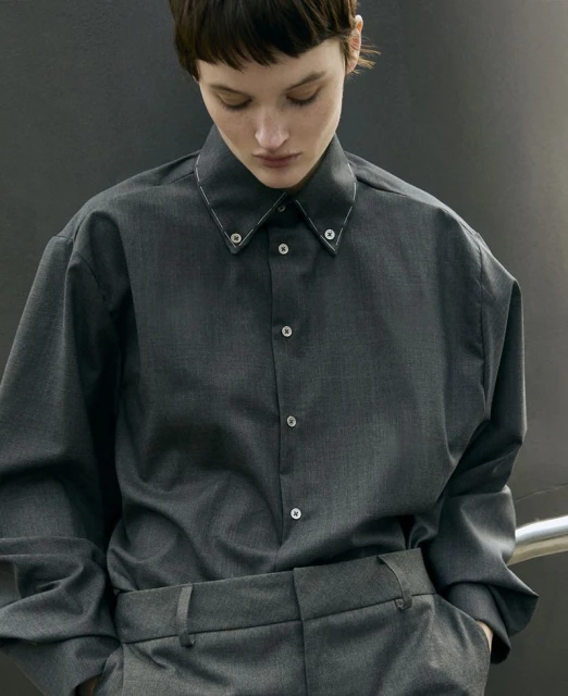 Portrait of a woman in a gray button-down shirt and matching trousers against a gray wall, looking down with a neutral expression.