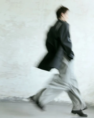 Motion blur photo of person walking fast in long coat and pants against a pale textured wall. Minimalist, contemporary style.