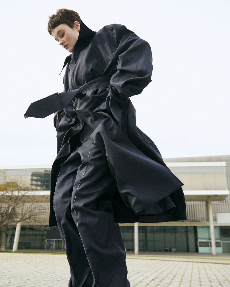 Model in a dark oversized belted trench coat and trousers standing outdoors against a modern building exterior.
