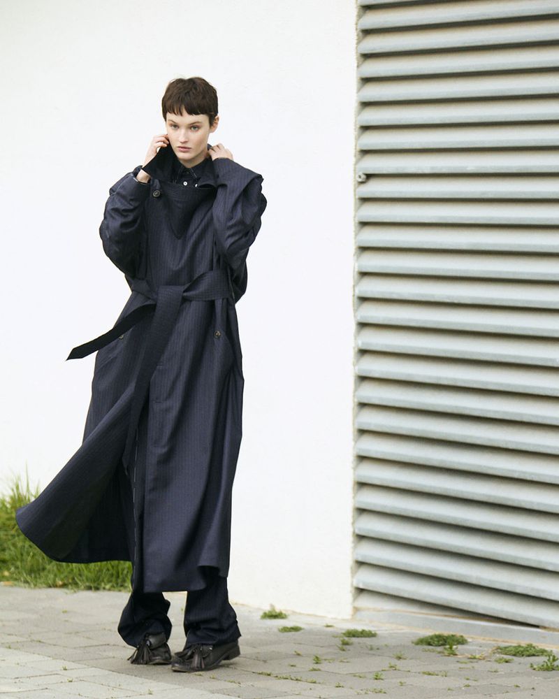 A woman with short dark hair wears a belted dark blue trench coat and matching pants, standing near a white wall and grey ventilation shaft.