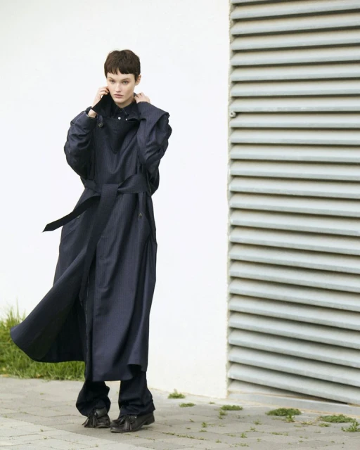 A woman with short dark hair wears a belted dark blue trench coat and matching pants, standing near a white wall and grey ventilation shaft.