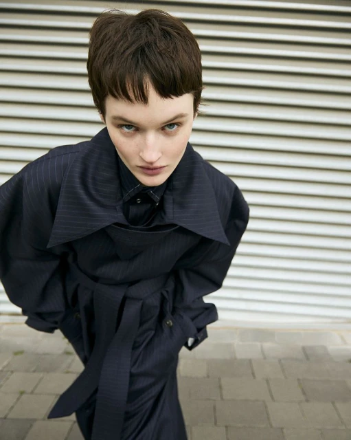A woman in a navy pinstripe trench coat poses against a white corrugated wall, shot from a high angle in an outdoor setting. 