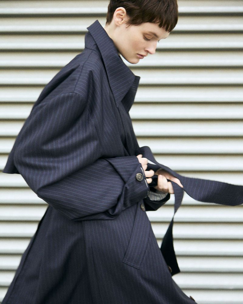 Model with short dark hair wearing a navy pinstripe trench coat with high collar against a slatted background