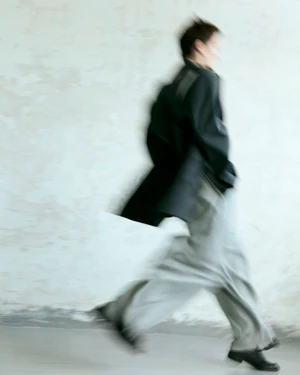 Motion blurred fashion editorial of a model wearing a dark jacket and loose trousers walking past a textured wall.