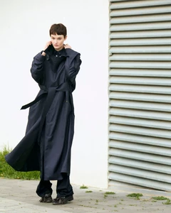 A model wearing a long dark blue belted trench coat posing outdoors against a white wall and a louvered metal shutter door.