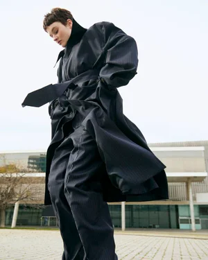 Model in a dark oversized belted trench coat and trousers standing outdoors against a modern building exterior.