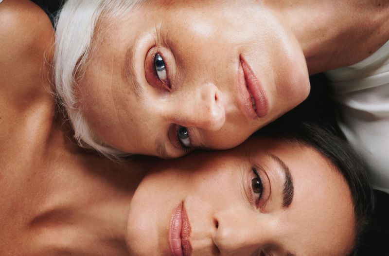 Close-up portrait of two adult females with fair skin, dark and light hair, lying closely together in a studio with white fabric.