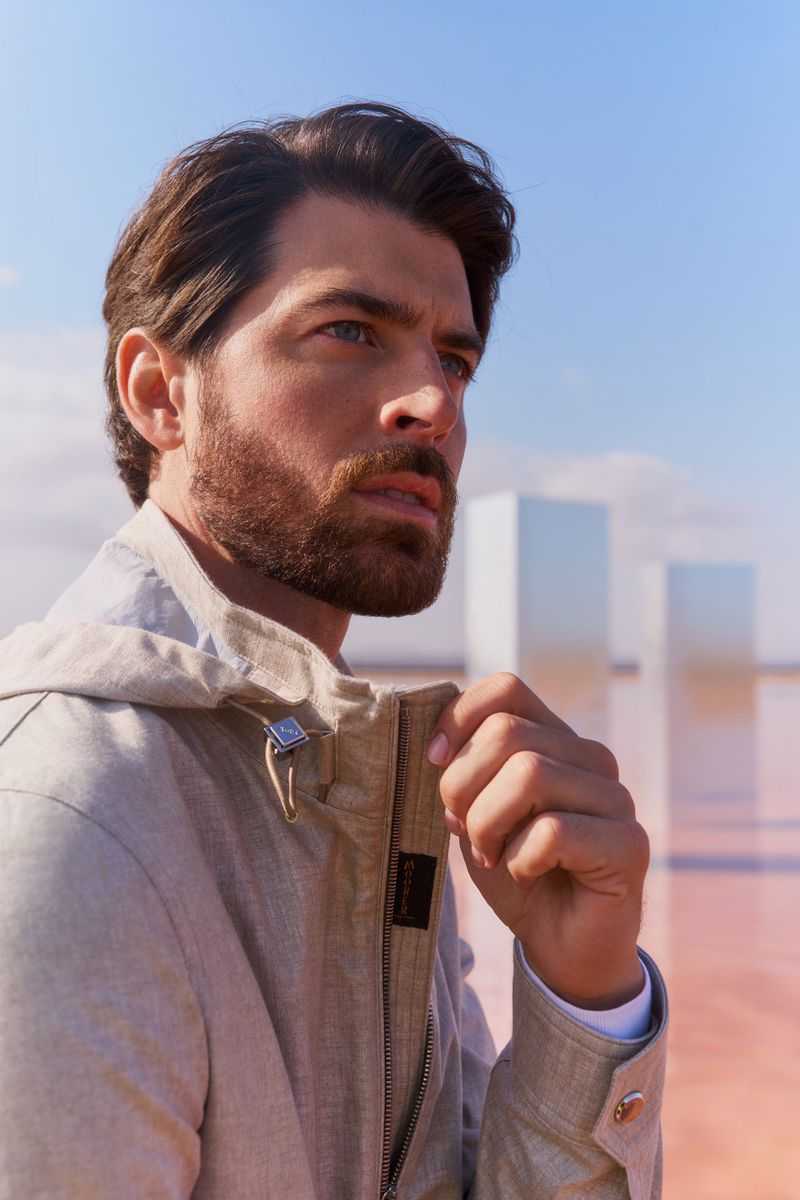 Portrait of an adult man with a beard wearing a hooded jacket outdoors against a blue sky with modern architecture in the distance.