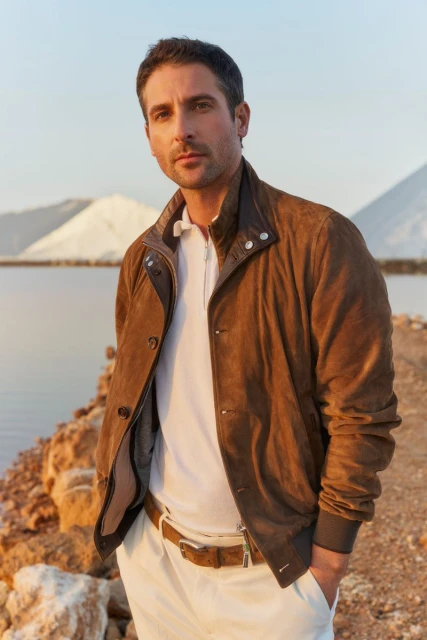 Portrait of a handsome man in a brown jacket and white pants outdoors, with a salt lake and piles of salt in the background.