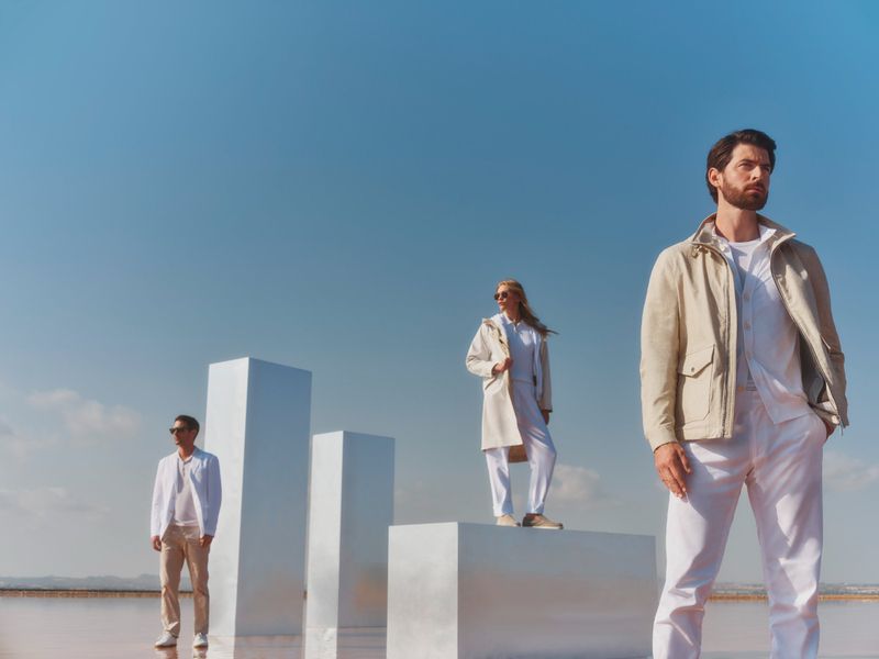 Three people in outdoor fashion shot. Two men and a woman standing in light-colored outfits, on white platforms, under a blue sky.