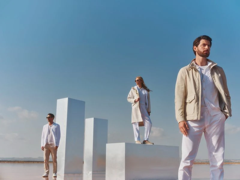 Three people in outdoor fashion shot. Two men and a woman standing in light-colored outfits, on white platforms, under a blue sky.