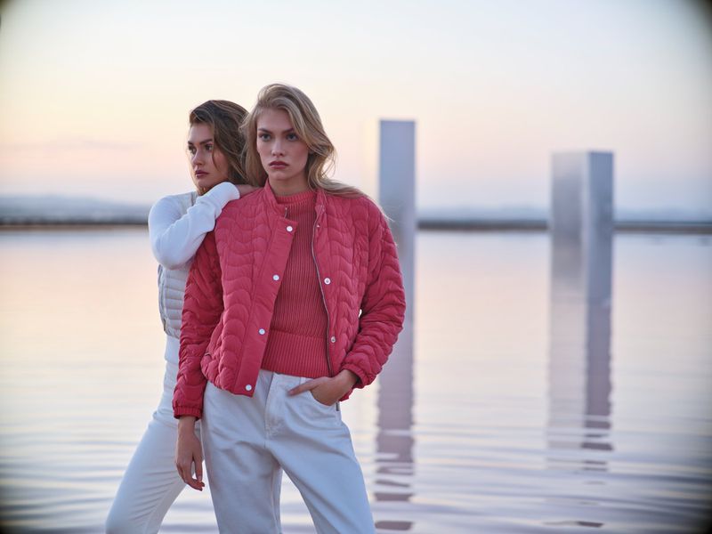 Two women in a fashion shoot in an outdoor setting, wearing stylish jackets and jeans with reflective art installations in the background.