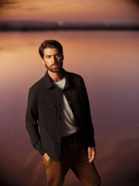 Portrait of a handsome man in a dark jacket and tan pants posing outdoors at sunset with blurred water in the background.