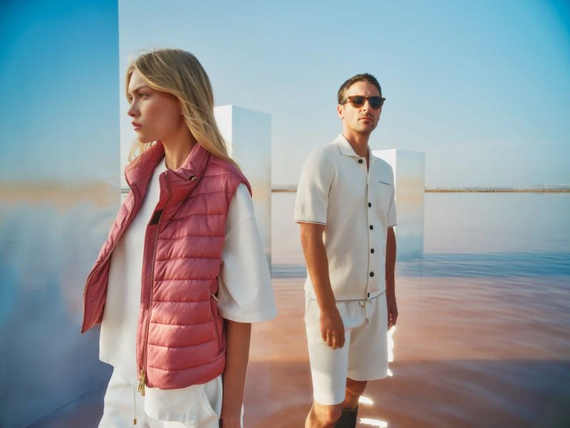 Blonde woman in a pink vest and a man with sunglasses pose in shallow, reflective water with mirrored cube in the background.