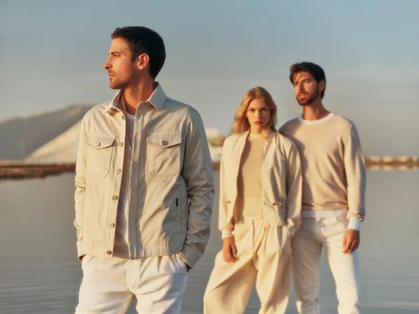 Three models in neutral beige and white apparel posing outdoors near calm water under a clear sky.