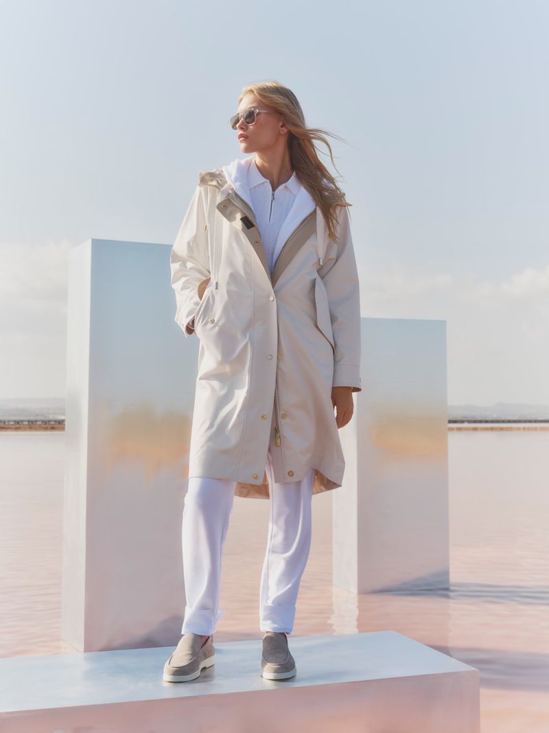 Fashionable blonde woman in a trench coat standing against sky and pink salt lake, accessorized with sunglasses and low-top shoes.
