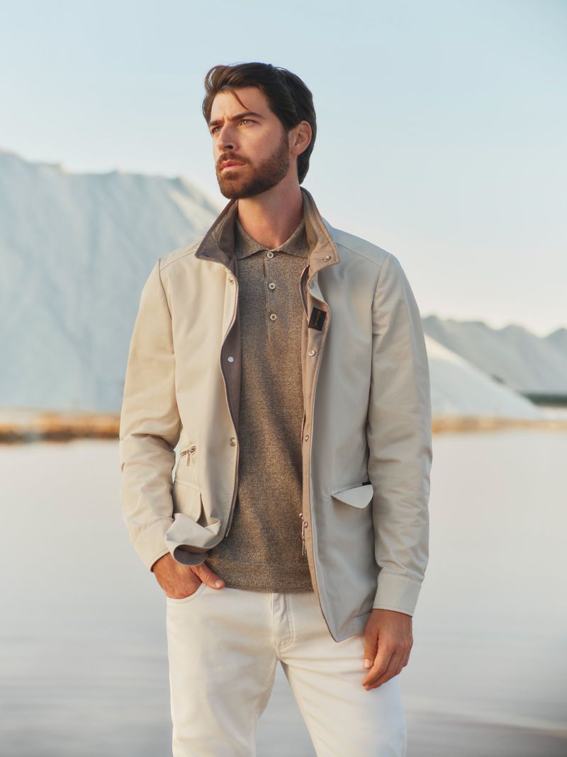 A handsome man with a beard in a light jacket and polo shirt stands outdoors against a bright, light blue sky and a backdrop of white mounds.