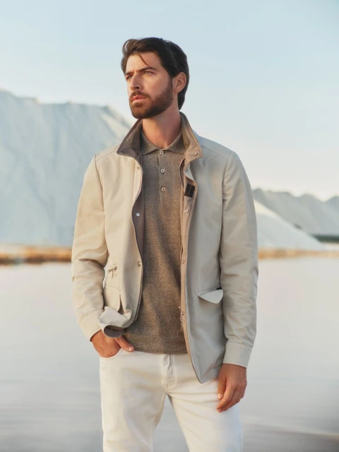 A handsome man with a beard in a light jacket and polo shirt stands outdoors against a bright, light blue sky and a backdrop of white mounds.