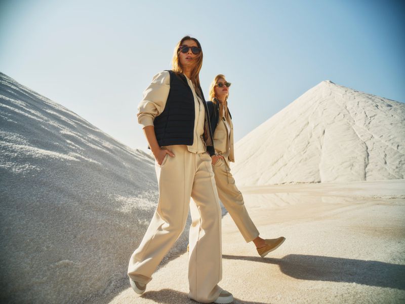Two women in casual streetwear walk on a sandy terrain under a blue sky. The woman in front wears a black vest. Fashion shot.
