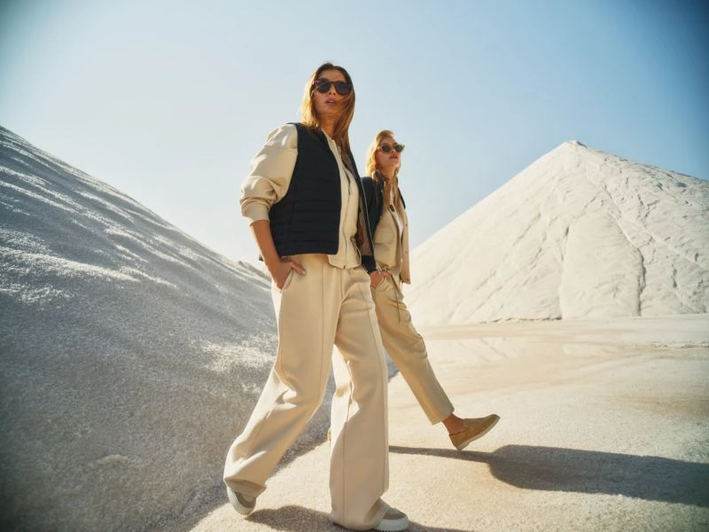 Two women in casual streetwear walk on a sandy terrain under a blue sky. The woman in front wears a black vest. Fashion shot.