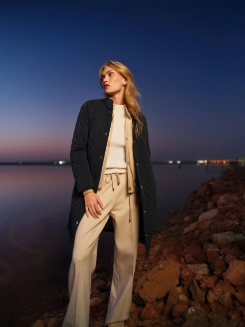 Fashionable blonde woman poses in a quilted jacket, sweater, and wide-leg pants against a twilight shoreline with a blurred cityscape.