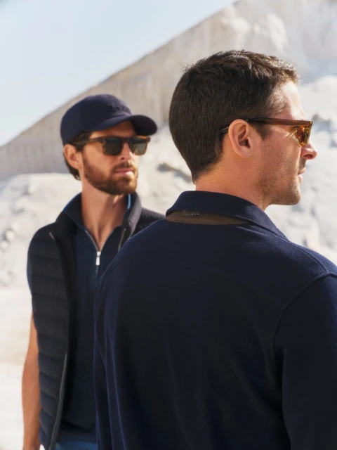 Two adult men in casual outfits and sunglasses stand against a bright white landscape. Fashion and style in an outdoor setting.