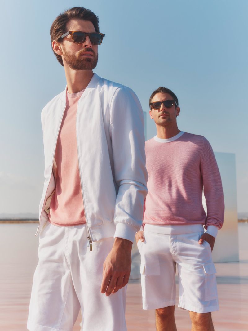 Two adult men stand in a light pink environment wearing sunglasses, pink shirts, and white shorts and jackets, posing in the sun.