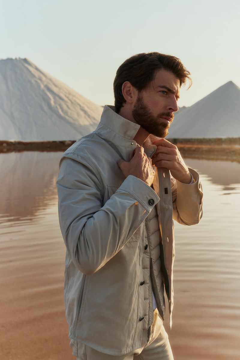 Bearded man in tan jacket poses in desert landscape with salt flats and pink water during golden hour in a fashion editorial.