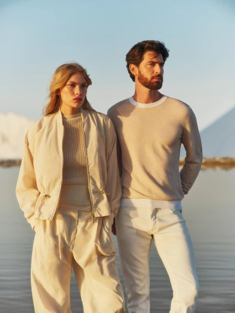 Fashion shot of a stylish young couple in matching sand-colored outfits posing in front of a body of water with mountains.