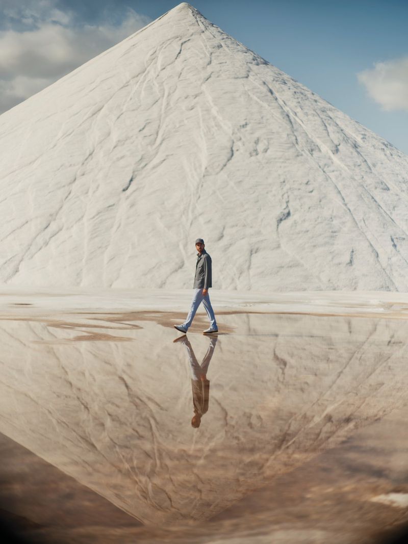 A man walks across a shallow reflective pool toward a large white mountain under a blue sky with soft clouds, creating a mirrored effect.