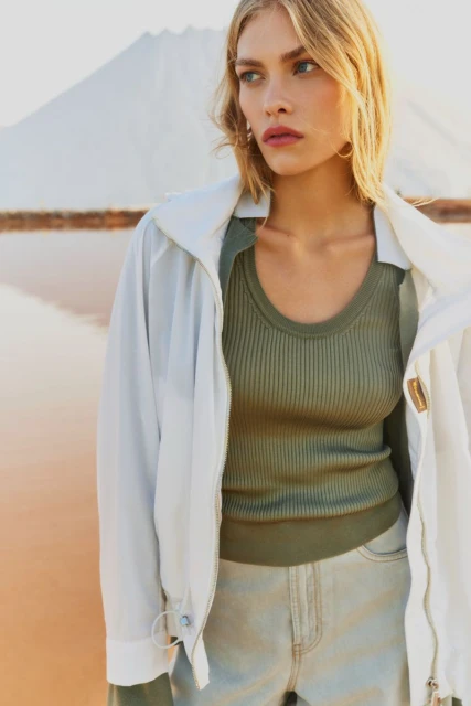 Close-up shot of a blonde woman in a jacket and jeans posing outdoors near a pink lake and white rock formation on a sunny day.