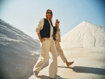 Two women wearing matching beige leisurewear and dark vests walking near large white sand dunes outdoors.