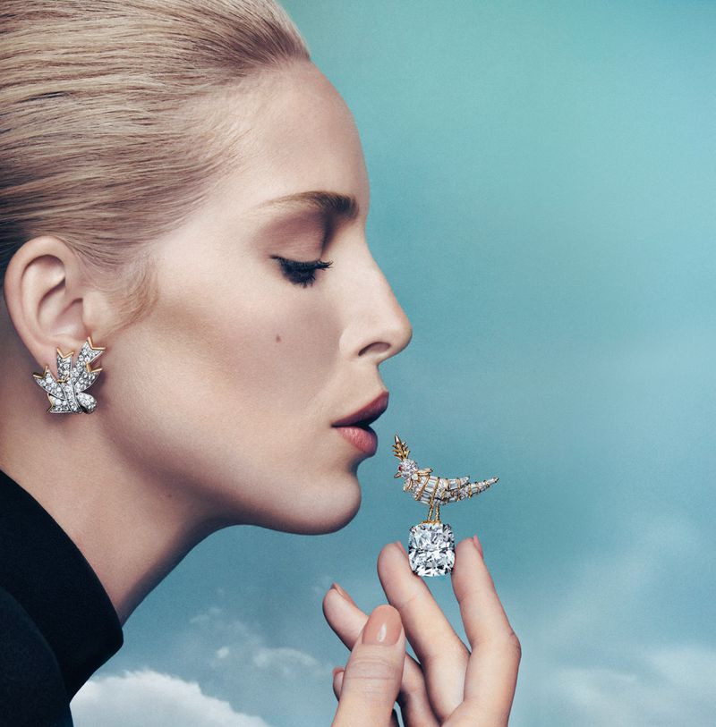 Elegant woman in profile wears diamond earrings and holds diamond bird jewelry against a blue gradient sky background in a fashion editorial.