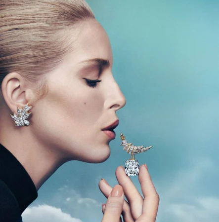 Elegant woman in profile wears diamond earrings and holds diamond bird jewelry against a blue gradient sky background in a fashion editorial.