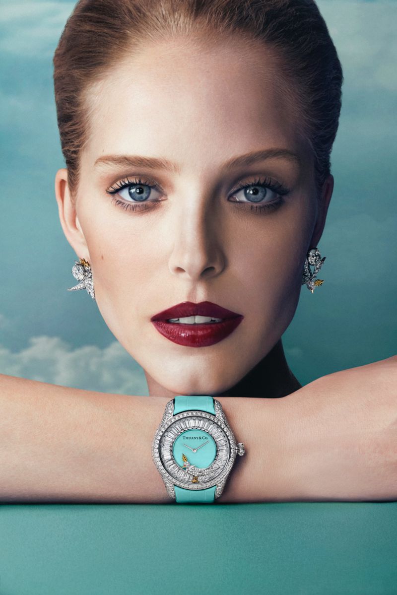 Close-up of a light-skinned woman with a Tiffany watch and earrings against a light blue, cloud-like background in a fashion editorial style.