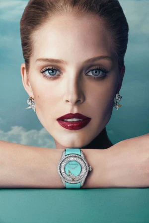 Close-up of a light-skinned woman with a Tiffany watch and earrings against a light blue, cloud-like background in a fashion editorial style.