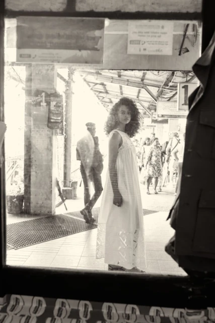 Woman in white dress stands at train platform, captured through window reflection, creating a vintage, atmospheric aesthetic.