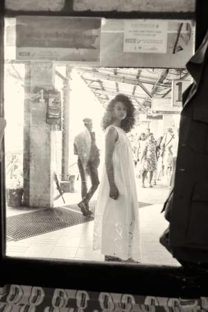 Woman in white dress stands at train platform, captured through window reflection, creating a vintage, atmospheric aesthetic.