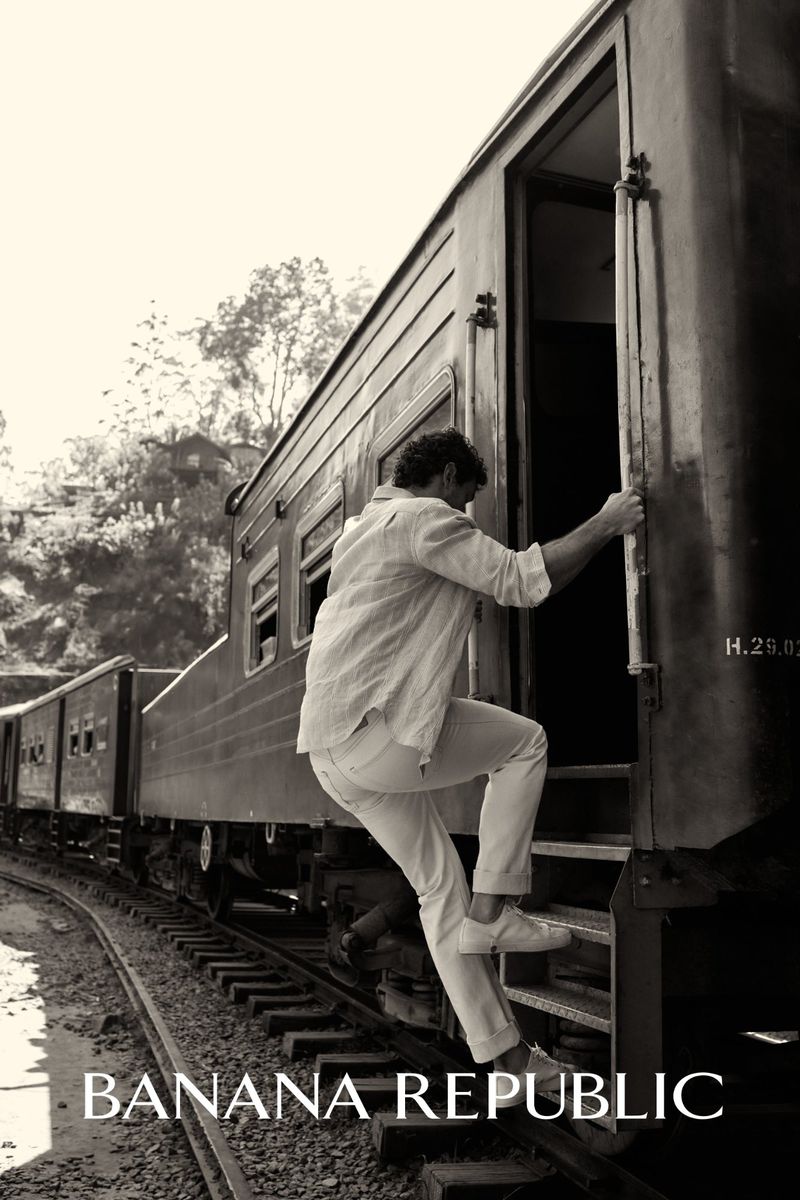 Man in white clothing steps onto a vintage train car with railroad tracks and trees visible in the monochrome background; Banana Republic logo.