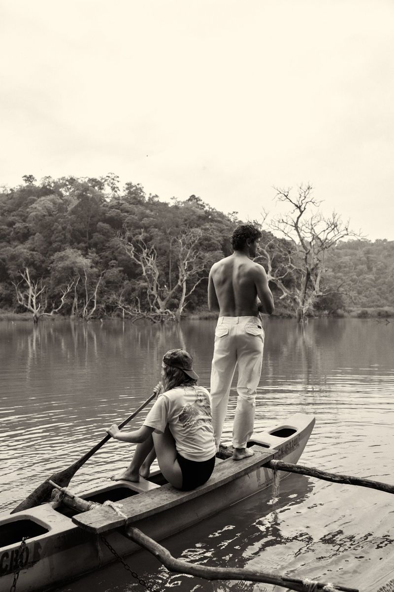 Monochrome image of two people in a dugout canoe on a lake surrounded by trees, one paddling and the other standing, facing the landscape.