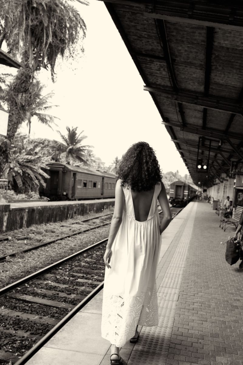Monochrome shot of a woman in a dress walking along train tracks with palm trees in the background at an outdoor train station.
